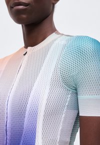 Gros plan d'une personne portant un maillot de cyclisme à manches courtes zippé avec un dégradé bleu, blanc et violet et une texture en maille.