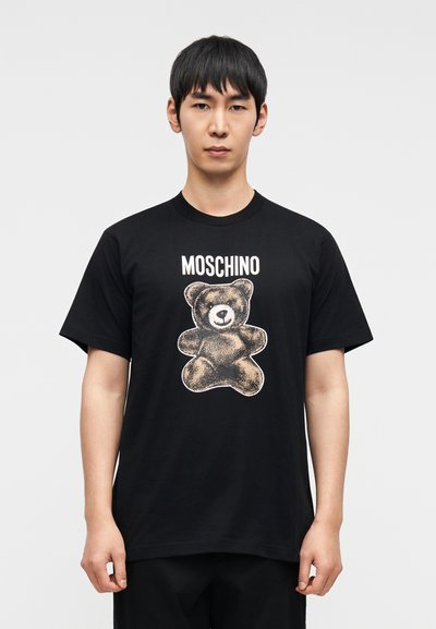 Μαύρο βαμβακερό t-shirt με γραφικό αρκούδας σε ανοιχτές αποχρώσεις και κείμενο "MOSCHINO" από πάνω. Χαλαρή εφαρμογή με κοντά μανίκια και στρογγυλή λαιμόκοψη.