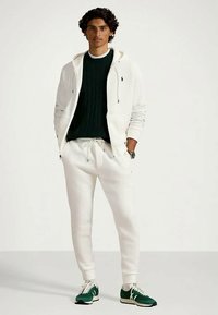 Młody mężczyzna stoi z rękami w kieszeniach białych dresów, ma na sobie białą bluzę z kapturem, ciemnozielony sweter oraz zielono-białe sneakersy.