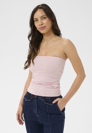 Donna con capelli lunghi e scuri che indossa un top rosa chiaro senza spalline e jeans blu scuro con la mano destra in tasca.