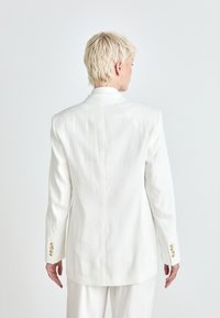 Blazer bianco sartoriale con spalline e polsini abbottonati, caratterizzato da un design posteriore pulito e una texture simile al lino.