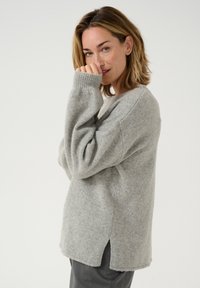 Pulover tricotat de culoare gri deschis, cu o croială relaxată, manșete ribelate, fante laterale și o textură moale. Designul are un guler rotund.