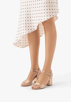 Nogi w sandałach na klockowym obcasie w kolorze różowego złota, z paskami wokół kostek, zestawione z białą sukienką w czarne groszki i asymetrycznym dołem.