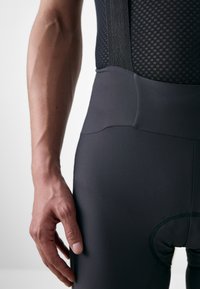 Schwarze Radsport-Bib-Shorts mit strukturiertem Mesh-Oberteil, enganliegendem Design, flachen Nähten und gepolstertem Sitzpolster für Komfort. Nahaufnahme einer Hand auf dem Kleidungsstück.