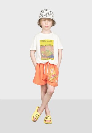 Vit T-shirt med en färgglad grafik, orange shorts med björntryck och gula tofflor med text. Grå bakgrund.