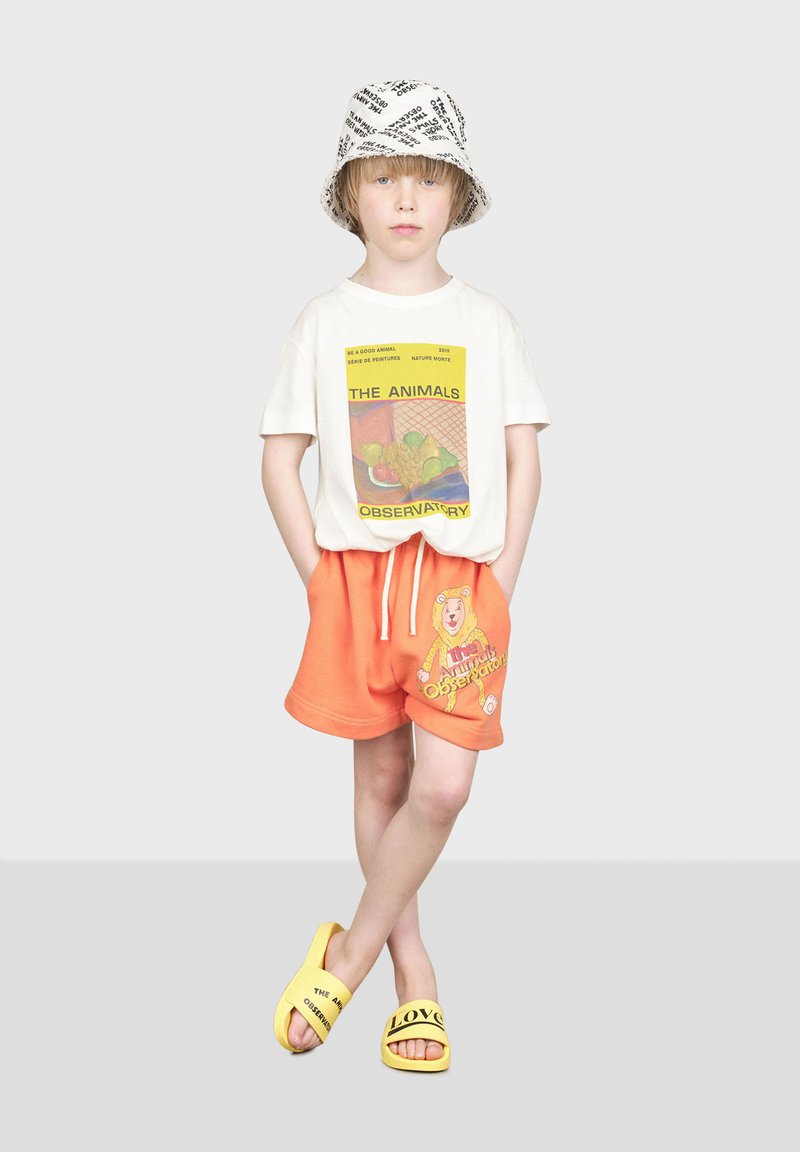 T-shirt blanche avec un graphique coloré, shorts orange avec un imprimé ours, et sandales jaunes avec du texte. Fond gris.