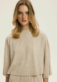 Mladá žena s blond vlasy po ramena, oblečená v béžovém žebrovaném topu s dlouhými rukávy, na jednoduchém světlém pozadí.