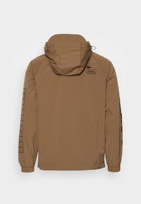 Brun huva jacka med elastiska ärmslut, med ett rutmönster på vänster ärm och logotyp på övre delen av ryggen. Slät, matt tyg.