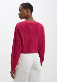 Maglione cropped in fucsia, con un motivo testurizzato e polsini a costine. Indossato con pantaloni bianchi a vita alta, che mostrano cuciture dettagliate.