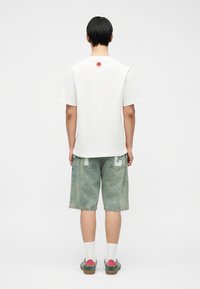 Άτομο που στέκεται με την πλάτη προς τα εμπρός, φοράει λευκό T-shirt με μικρό κόκκινο λογότυπο στο πάνω μέρος της πλάτης, σορτς από τζιν με λευκά γράμματα, λευκές κάλτσες και πολύχρωμα αθλητικά παπούτσια.