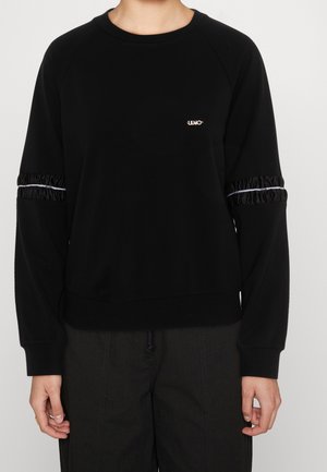 Personne portant un sweat-shirt à manches longues noir avec des bandes de tissu froncées et une bordure blanche sur le haut des manches ainsi qu'un petit logo blanc sur la poitrine.