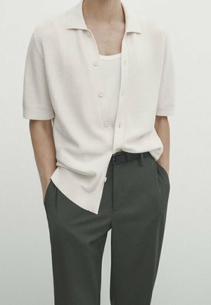 Homme portant un cardigan boutonné à manches courtes crème par-dessus un débardeur blanc, avec un pantalon vert foncé et les mains dans les poches.
