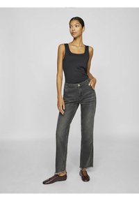 Top negro sin mangas combinado con jeans gris de cintura alta con dobladillo deshilachado y bolsillos. Modelo llevando zapatos marrones. Fondo neutro.