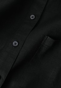 Gros plan sur une chemise en tissu noir montrant deux boutons noirs avec le texte "TRUTH" et une poche poitrine sur un matériau en lin texturé.