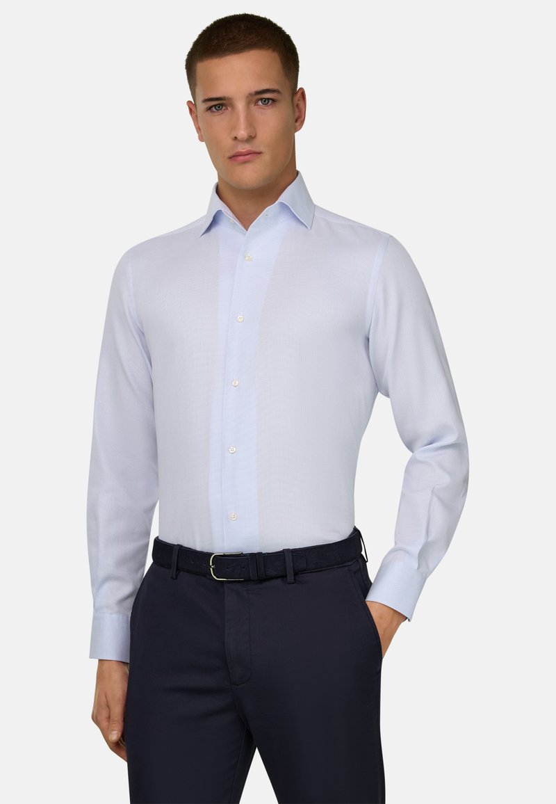 Homme portant une chemise habillée à manches longues bleu clair rentrée dans un pantalon foncé avec ceinture noire, debout avec une main dans la poche.
