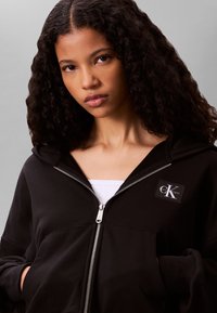 Giovane donna con capelli ricci che indossa una felpa nera con cerniera e una maglietta bianca sotto, su uno sfondo grigio uniforme.