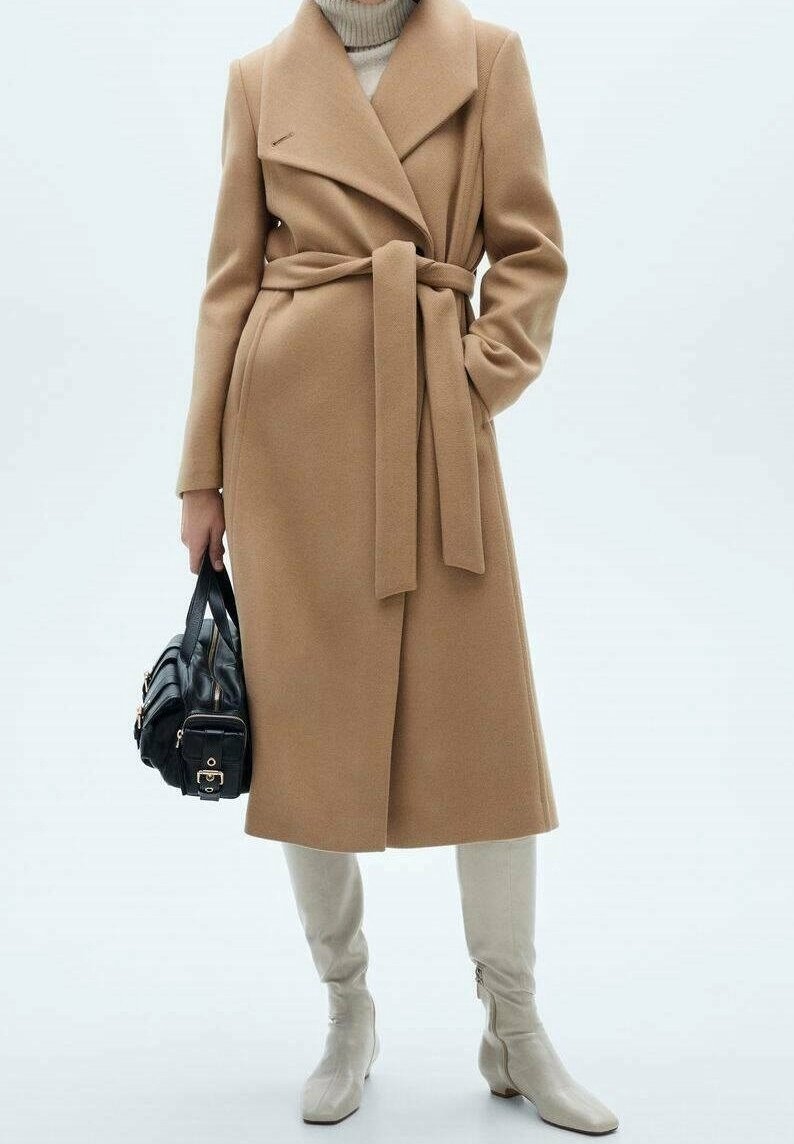 Kabát z kamelíkové vlněné směsi s zavinovacím designem, širokými klopami a páskem na zavázání, doplněný béžovými kozačkami po kolena a černou kabelkou.