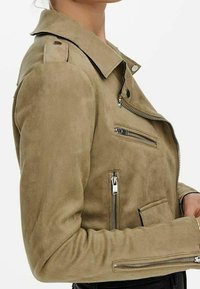 Kvinna som bär en beige mockabikerjacka med dragkedjedetaljer på bröstet, ärmarna och fickan, visad från sidan upp till halsen.