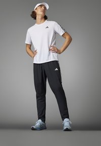 Weißes Sport-T-Shirt mit schwarzen drei Streifen Akzenten, kombiniert mit schwarzen Jogginghosen. Hellblaue Laufschuhe ergänzen das Outfit.