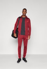 Homme en survêtement de sport rouge et baskets noires, portant un sac de sport noir sur l'épaule, se tenant devant un fond uni clair.