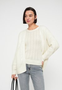 Νεαρή γυναίκα με κοντά σκούρα μαλλιά, που φοράει λευκό πλεκτό πουλόβερ και ζακέτα, κρατώντας μια μαύρη τσάντα χειρός, στέκεται μπροστά σε απλό φόντο.