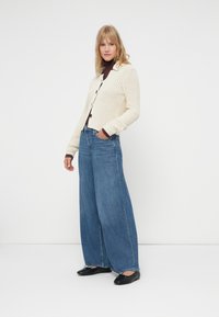 Cárdigan de punto crema con botones negros, usado sobre una blusa marrón, combinado con unos pantalones vaqueros de pernera ancha azul y bailarinas negras.