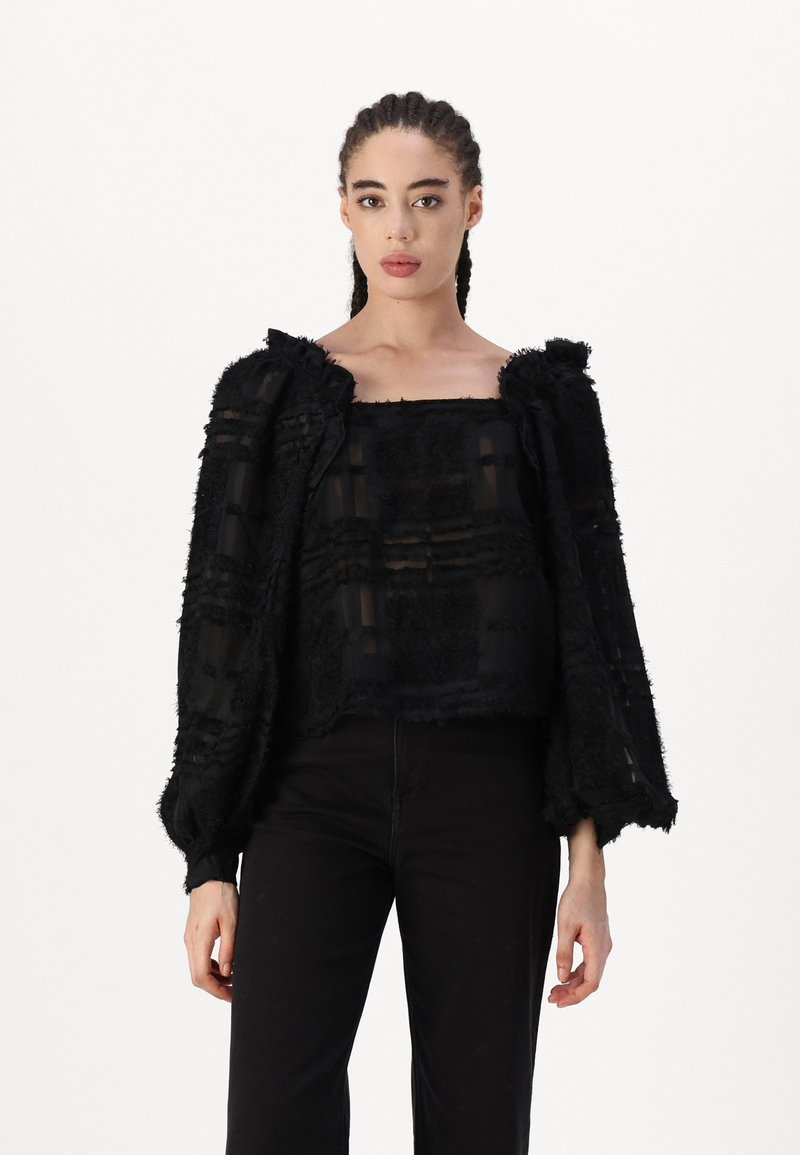 Schwarzes, strukturiertes Cropped-Top mit transparenten Abschnitten, weiten Ärmeln und fransigen Kanten. Quadrat-Ausschnitt mit einer kastigen Passform. Kombiniert mit schwarzen Hosen.