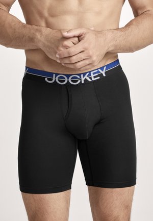 Muscular man wearing black Jockey boxer briefs with blue waistband, standing with hands clasped in front of bare torso.