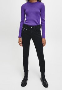 Suéter morado con acanalado y mangas largas, combinado con jeans ajustados negros de tiro alto que cuentan con bolsillos delanteros con cremallera y botas negras con cordones.
