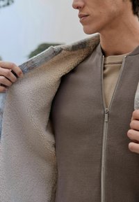 Bunda na zips so mäkkou textúrovanou vnútornou podšívkou, v neutrálnom taupe odtieni. Vonkajšia vrstva má jemný vzorovaný golier.