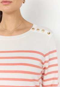 Mujer vistiendo un suéter a rayas blancas y coral con botones dorados en el hombro, aro dorado en la oreja y delicada cadena dorada con un pequeño colgante.