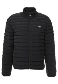 Svart kviltad pufferjacka med hög krage och dragkedja framtill, med en liten grön Lacoste-logotyp på vänstra bröstområdet.