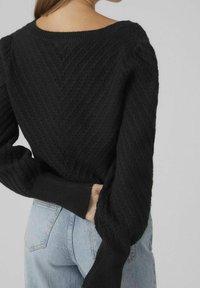 Maglione nero a maglia con un motivo a chevron testurizzato, scollatura ampia e polsini aderenti, abbinato a jeans blu chiaro.