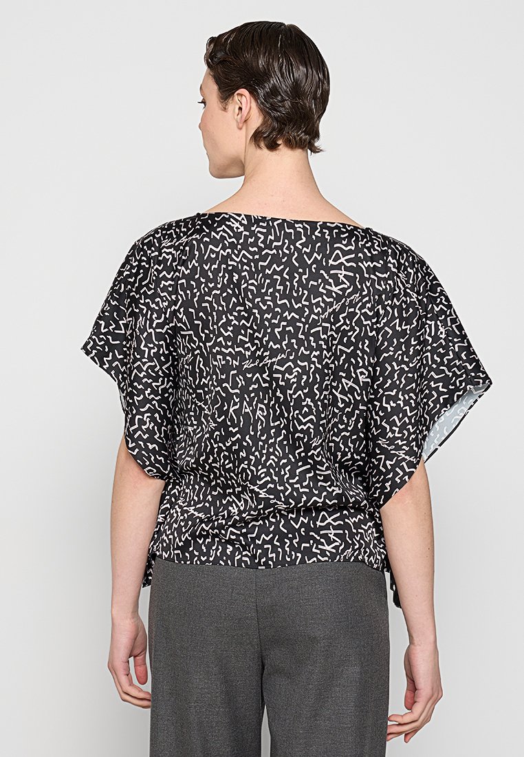 Homme portant une chemise à manches courtes avec un motif abstrait noir et blanc et un pantalon gris, vu de dos sur un fond uni.