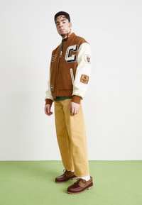 Veste de varsity marron avec des manches crème, grand patch "C", pantalon beige, et chaussures marron avec une semelle texturée, sur un fond clair.