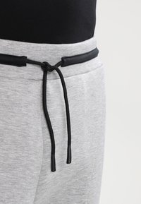 Grå sweatpants har en svart snörning med plastspetsar och en platt svart midja. Tyget har en slät textur och en avslappnad design.