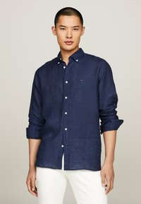 Camicia in lino blu navy con colletto button-down. Bottoni bianchi e un piccolo logo sul petto. Maniche lunghe arrotolate ai polsi.