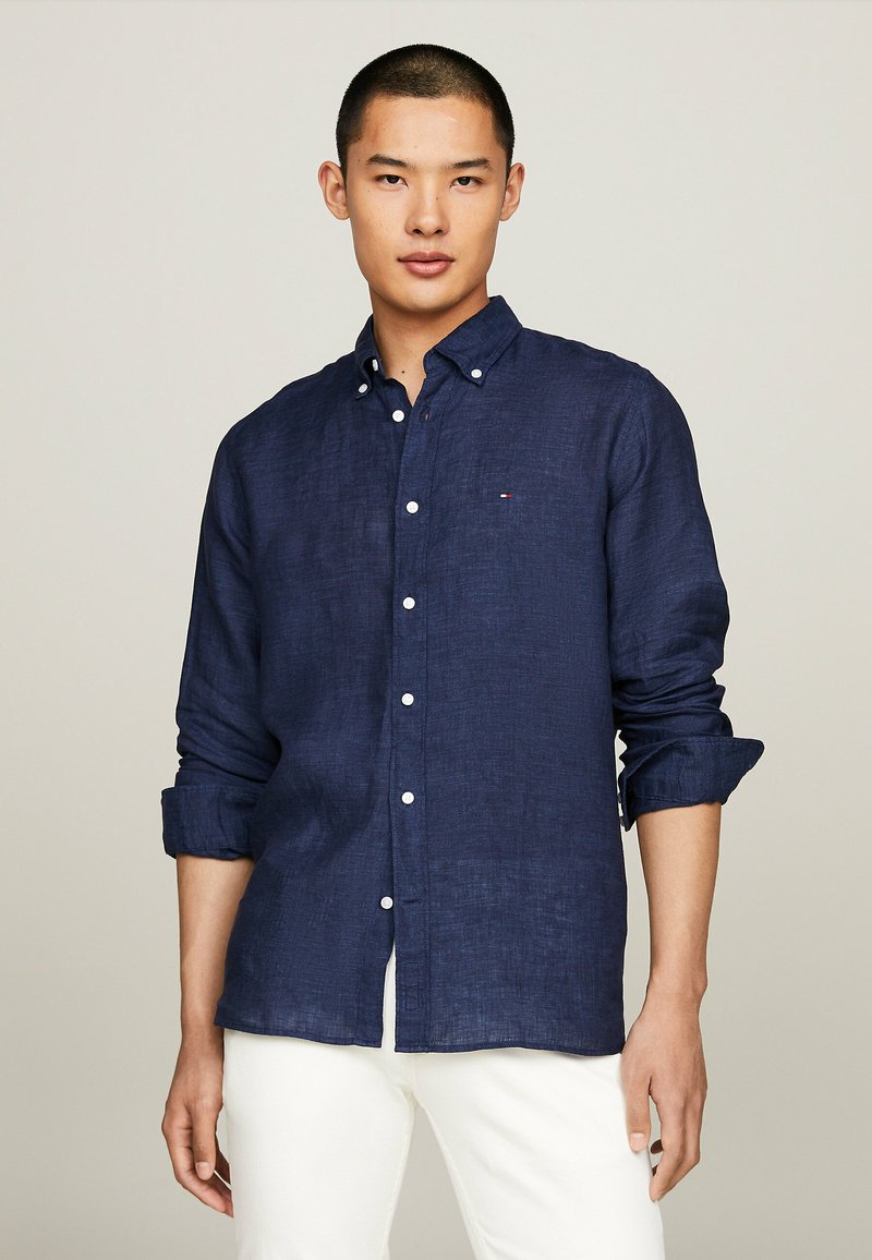 Camicia in lino blu navy con colletto button-down. Bottoni bianchi e un piccolo logo sul petto. Maniche lunghe arrotolate ai polsi.