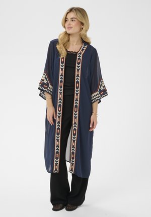 Femme portant un kimono bleu marine ouvert à l'avant avec une broderie géométrique sur une tenue noire, debout devant un fond blanc uni.