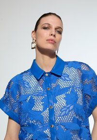 Blue shirt featuring a floral lace design, short sleeves, and a collared neckline with gold buttons. Textured fabric and embroidered accents.