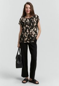 Sort og brun mønstret top, afslappet pasform, korte ærmer, kombineret med sorte bukser med lige ben og sorte sandaler, der holder en sort håndtaske.