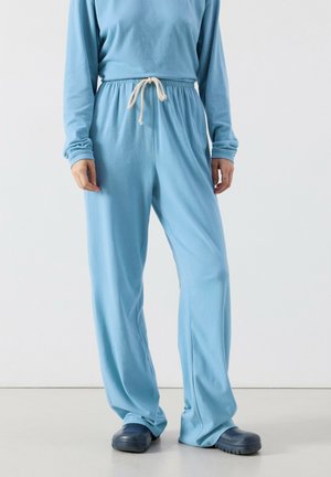 Person trägt locker sitzende hellblaue Hosen mit Kordelzugbund und dunkelblaue Slip-On-Schuhe, steht vor einem einfarbigen Hintergrund.