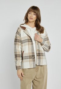 Beżowa, brązowa i kremowa koszula w kratę, wykonana z teksturowanej tkaniny, z kapturem i brązowymi skórzanymi detalami. Noszona na białym hoodie.