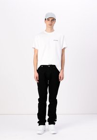 Camiseta blanca con un pequeño logotipo de texto, jeans negros y zapatillas blancas. La gorra es de color azul claro. El conjunto tiene un aspecto casual con un ajuste relajado.