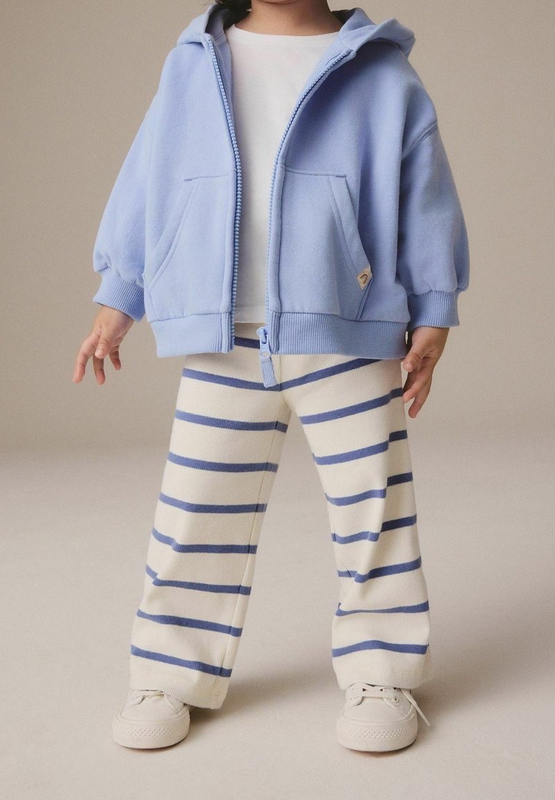 Enfant portant un sweat à capuche bleu clair zippé par-dessus un t-shirt blanc, un pantalon crème avec des rayures bleues, et des baskets blanches, debout sur un fond neutre.