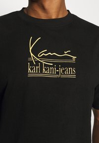 Camiseta negra de algodón con cuello redondo. Presenta un logo impreso en dorado y el texto "karl kani-jeans" en la parte frontal.