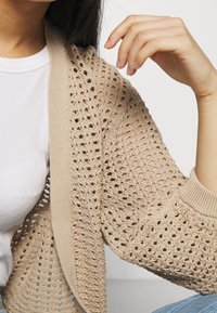 Beige neulottu neuletakki, jossa on pitsikuviollinen kuvio, resorit hihansuuissa ja pyöreä pääntie, käytetty valkoisen paidan päällä. Näkyvä rakenne ja kevyt kangas.