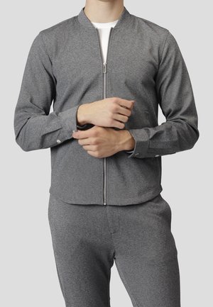 Man wearing a gray zip-up jacket and matching gray pants, adjusting the cuff of the left sleeve over a white shirt.