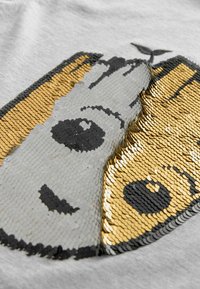 Grauer Stoff mit einem strukturierten Paillettendesign, das schwarze, goldene und silberne Pailletten zeigt und ein stilisiertes Gesicht mit Blattdetail darstellt.