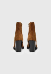 Brune suède ankelstøvler med en solid sort blokk hæl. Har en sidelukning med glidelås og en glatt tekstur; minimalistisk design, ingen synlige mønstre.